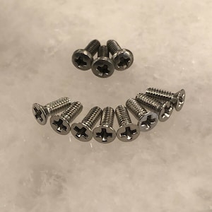 May include: A close-up of a group of silver screws with a cross-shaped head. The screws are arranged in a semi-circle on a white surface.