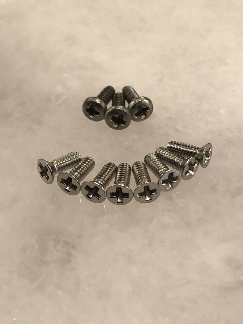 May include: A close-up of a group of silver screws with a cross-shaped head. The screws are arranged in a semi-circle on a white surface.