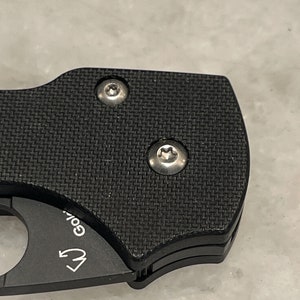 K&ouml;nnte beinhalten: Nahaufnahme eines schwarzen Taschenmessers mit einem strukturierten Griff. Zwei silberne Schrauben sind am Griff sichtbar.