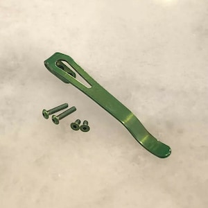 May include: A green metal pocket clip with four small screws. The clip is curved and has a small opening at the top.