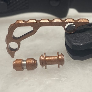 May include: A set of three copper-colored metal accessories for a pocket knife. The largest piece is a hook-shaped tool with a serrated edge. The two smaller pieces are cylindrical and have a textured surface.