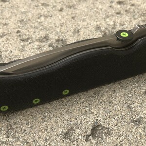 May include: A black folding knife with a green accent on the handle and a silver blade. The knife has a textured handle and a small green screw on the side.
