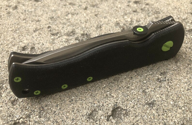 May include: A black folding knife with a green accent on the handle and a silver blade. The knife has a textured handle and a small green screw on the side.