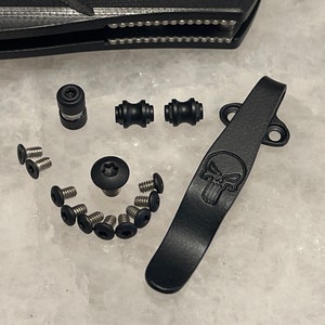 May include: A black pocket clip with a skull design, a black screw, two black cylindrical spacers, and a set of small silver screws.