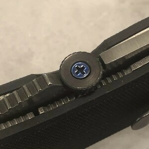 May include: Close-up of a black folding knife with a silver blade and a textured handle. The knife features a blue screw and a silver metal clip. The blade is partially open, revealing the serrated edge.