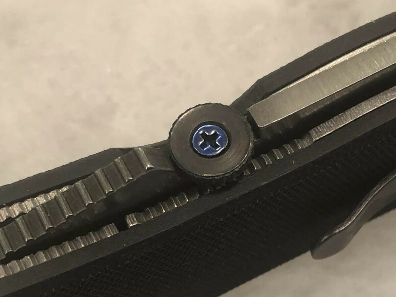 May include: Close-up of a black folding knife with a silver blade and a textured handle. The knife features a blue screw and a silver metal clip. The blade is partially open, revealing the serrated edge.
