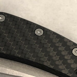 以下が含まれることがあります： 黒いカーボンファイバー製のナイフのハンドルをクローズアップ。銀色のネジが3つ付いている。