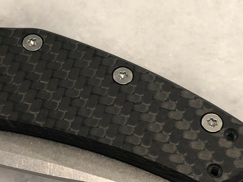 以下が含まれることがあります： 黒いカーボンファイバー製のナイフのハンドルをクローズアップ。銀色のネジが3つ付いている。