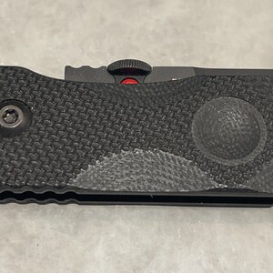 May include: Black folding knife with a textured black handle and red accents. The knife has a partially visible blade and a small red button on the handle.