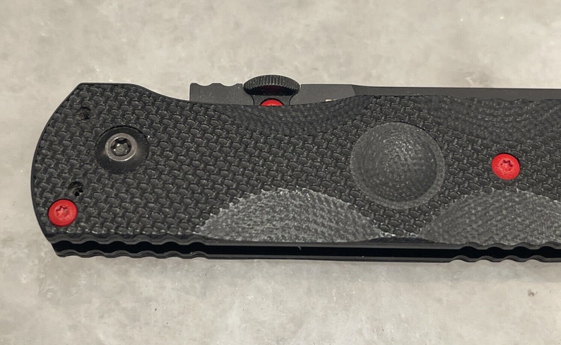 May include: Black folding knife with a textured black handle and red accents. The knife has a partially visible blade and a small red button on the handle.