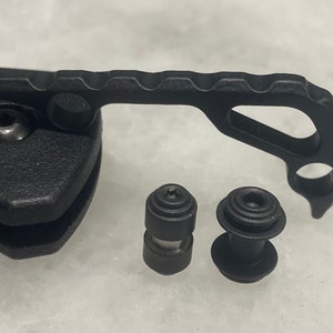 May include: Close-up of a black knife handle with a textured grip and a black metal hook. Two black metal screws are visible in the foreground.