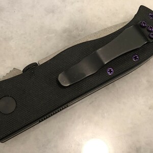 May include: A black pocket knife with a black handle and a silver blade. The knife has a purple clip and purple screws.