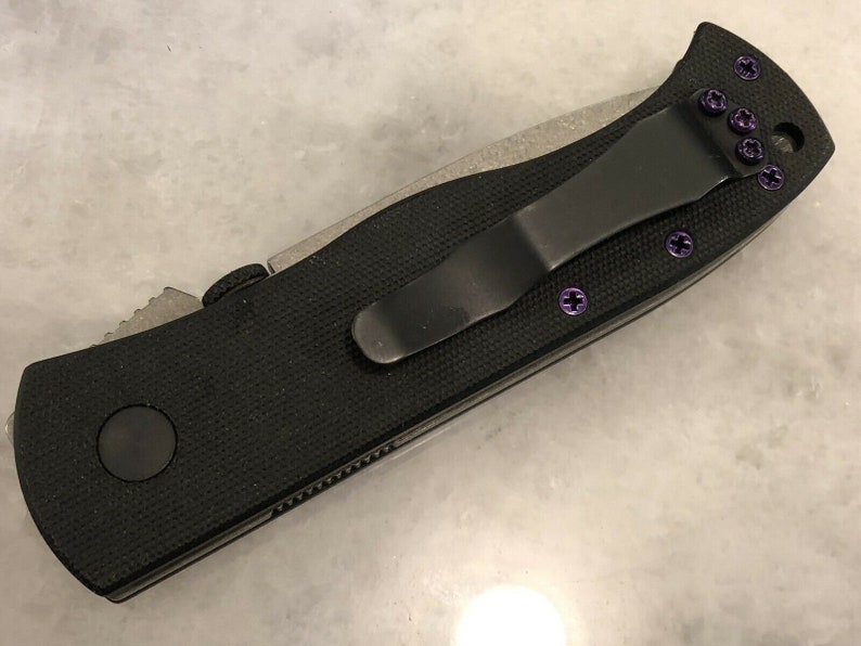May include: A black pocket knife with a black handle and a silver blade. The knife has a purple clip and purple screws.