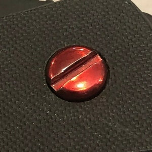 May include: A close-up of a red screw with a single slot on a black textured surface.