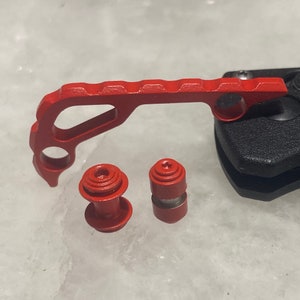 May include: Red metal knife handle with two red metal screws. The handle has a textured surface and a hole in the center.