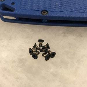 May include: A close-up of a pile of small black screws. The screws are arranged in a loose pile on a white surface.