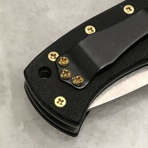 May include: Close-up of a black pocket knife with a gold clip and gold screws. The knife has a black handle and a silver blade.