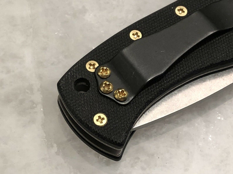 May include: Close-up of a black pocket knife with a gold clip and gold screws. The knife has a black handle and a silver blade.