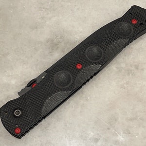 May include: Black folding knife with a textured handle and red accents. The handle has a textured pattern and four circular indents. The knife has a black blade and a silver clip.