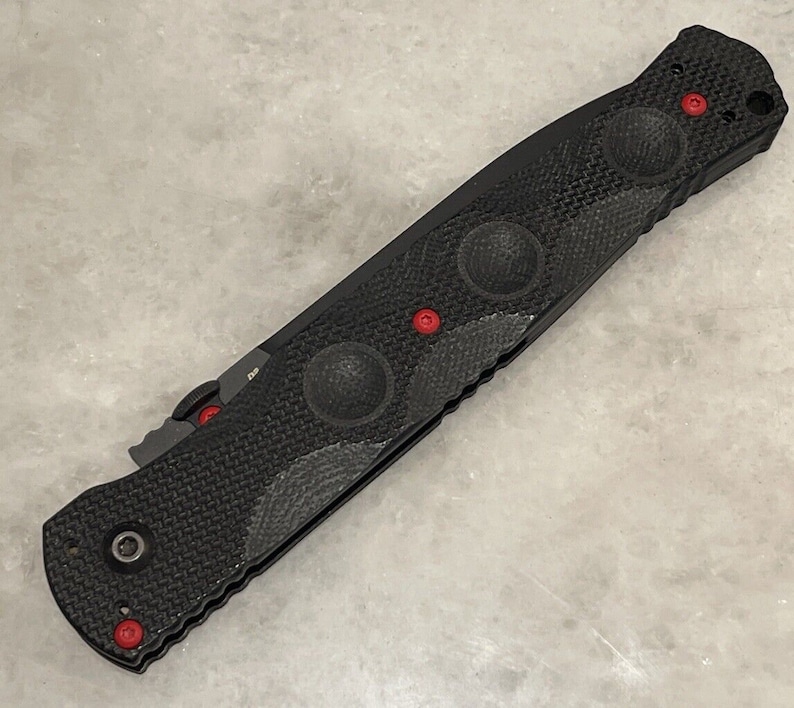 May include: Black folding knife with a textured handle and red accents. The handle has a textured pattern and four circular indents. The knife has a black blade and a silver clip.
