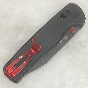 Pu&ograve; includere: Un coltello pieghevole nero con clip da tasca e dettagli rossi. Il coltello ha un'impugnatura strutturata e una lama parzialmente visibile. Il testo "USA" e "6105" &egrave; stampato sulla lama. Il coltello &egrave; mostrato su una superficie grigio chiaro.
