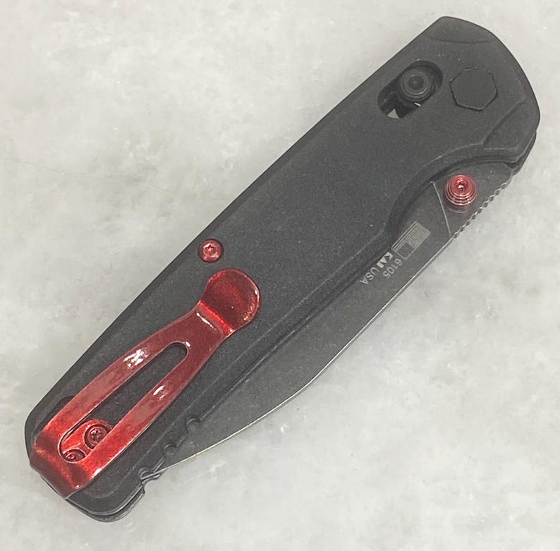 Pu&ograve; includere: Un coltello pieghevole nero con clip da tasca e dettagli rossi. Il coltello ha un'impugnatura strutturata e una lama parzialmente visibile. Il testo "USA" e "6105" &egrave; stampato sulla lama. Il coltello &egrave; mostrato su una superficie grigio chiaro.