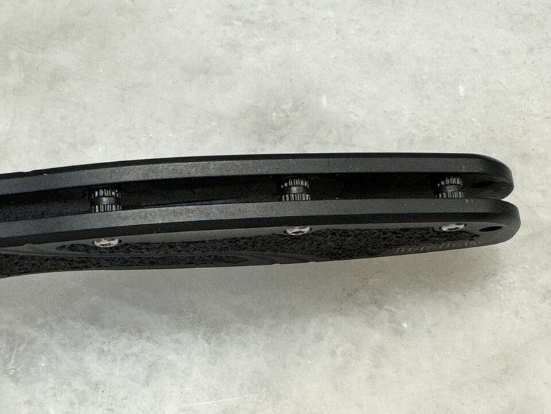 May include: Close-up of a black folding knife with a textured handle and three visible screws.