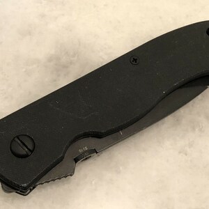 May include: A black folding knife with a textured handle and a partially open blade. The blade has a serrated edge and a small inscription that reads "S75".