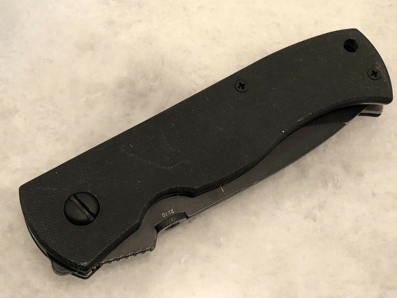 May include: A black folding knife with a textured handle and a partially open blade. The blade has a serrated edge and a small inscription that reads "S75".