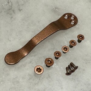 May include: A copper-colored titanium handle with a rounded end and five small screws. The handle has a textured surface and three holes on the top.
