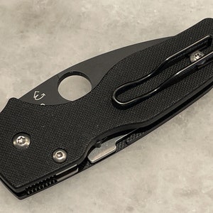 K&ouml;nnte beinhalten: Schwarzes Taschenmesser mit schwarzem Griff und silberner Klinge. Das Messer hat einen Taschenclip und ein kleines Loch im Griff.