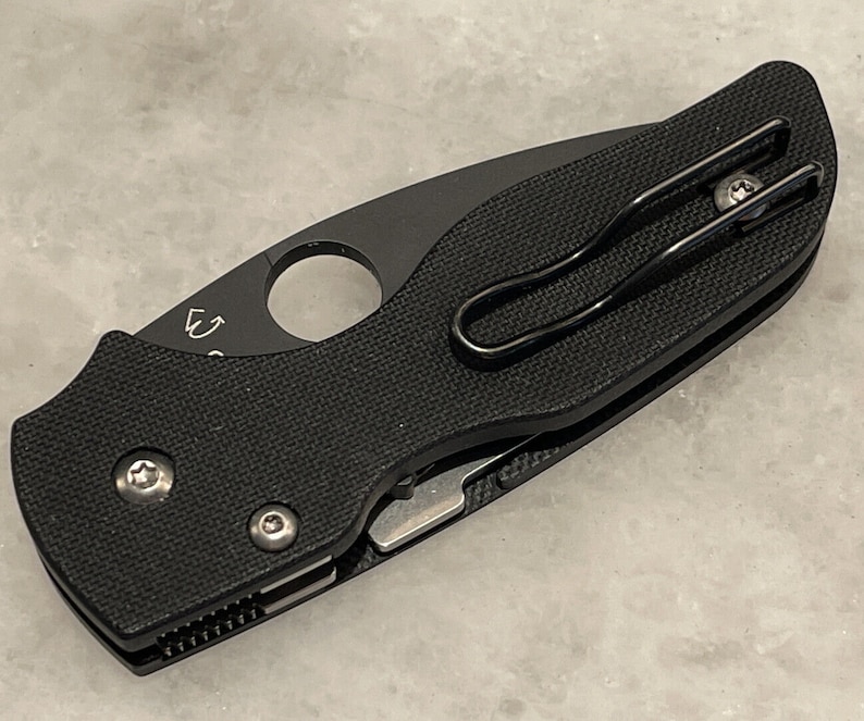K&ouml;nnte beinhalten: Schwarzes Taschenmesser mit schwarzem Griff und silberner Klinge. Das Messer hat einen Taschenclip und ein kleines Loch im Griff.