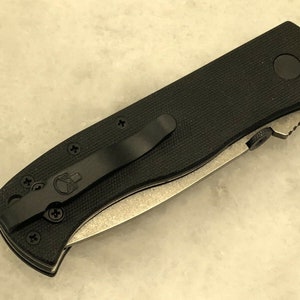 May include: Black folding knife with a black handle and a silver blade. The handle has a skull logo and a pocket clip.