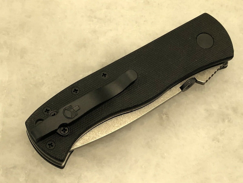 May include: Black folding knife with a black handle and a silver blade. The handle has a skull logo and a pocket clip.
