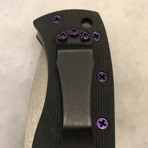 May include: A black pocket knife with a black clip and purple screws. The knife has a textured handle and a silver blade.