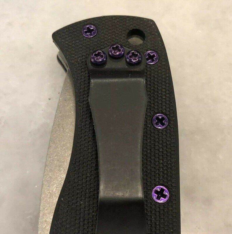 May include: A black pocket knife with a black clip and purple screws. The knife has a textured handle and a silver blade.