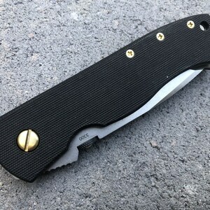 May include: A black folding knife with a black handle and a silver blade. The handle has gold screws and a gold accent on the side. The blade is partially open and the knife is lying on a gray surface.