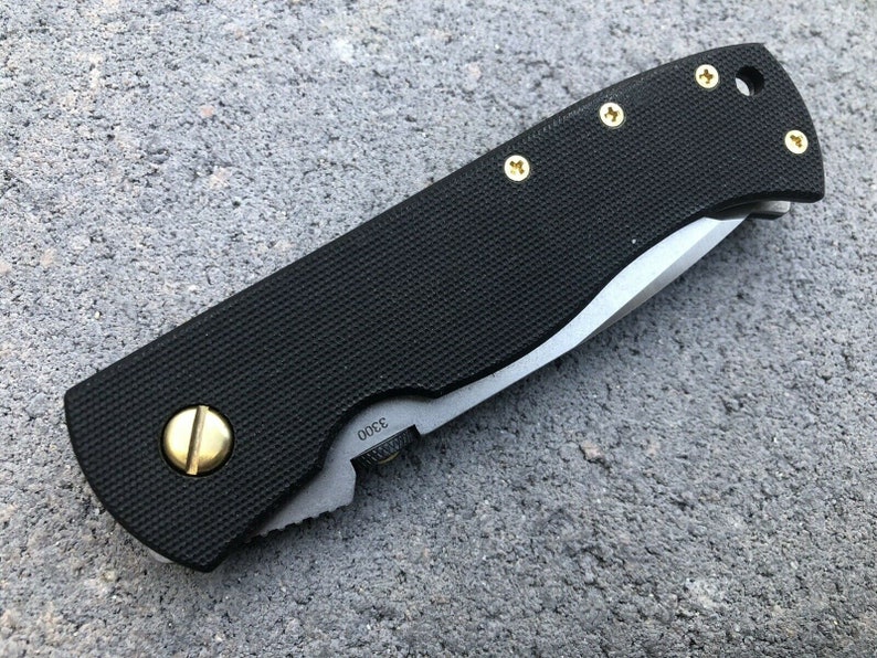 May include: A black folding knife with a black handle and a silver blade. The handle has gold screws and a gold accent on the side. The blade is partially open and the knife is lying on a gray surface.