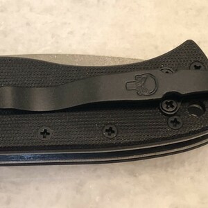 May include: Black pocket knife with a black pocket clip and a skull logo. The knife has a textured black handle and a silver blade.