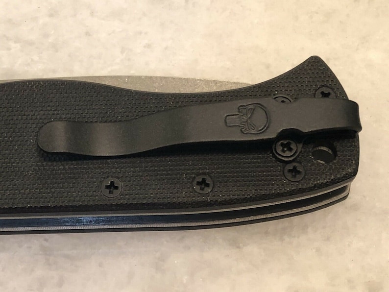 May include: Black pocket knife with a black pocket clip and a skull logo. The knife has a textured black handle and a silver blade.