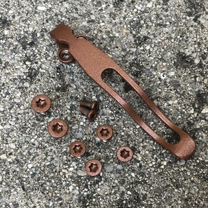 May include: A copper-colored pocket clip with six copper-colored screws. The pocket clip is designed to attach to a pocket knife or other tool.