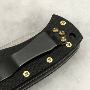 May include: Close-up of a black pocket knife with a gold clip and gold screws. The knife has a black handle and a silver blade.