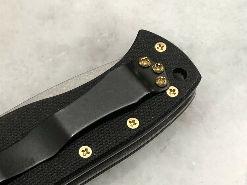 May include: Close-up of a black pocket knife with a gold clip and gold screws. The knife has a black handle and a silver blade.