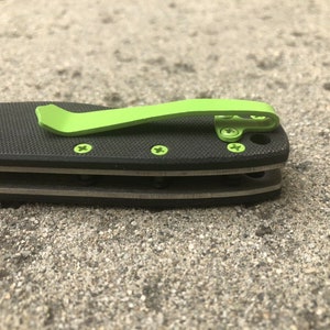 Pode incluir: Uma faca dobr&aacute;vel preta com um clipe de bolso verde. A faca tem uma al&ccedil;a texturizada e uma l&acirc;mina prateada.