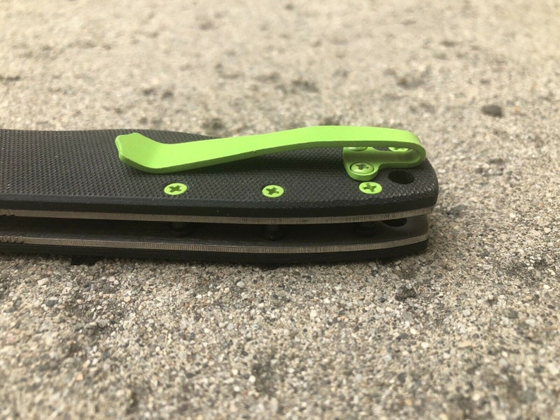 Pode incluir: Uma faca dobr&aacute;vel preta com um clipe de bolso verde. A faca tem uma al&ccedil;a texturizada e uma l&acirc;mina prateada.