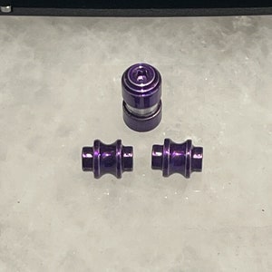 Puede incluir: Tres cuentas de metal morado, dos son cilíndricas y una es redonda con una ranura alrededor del medio.