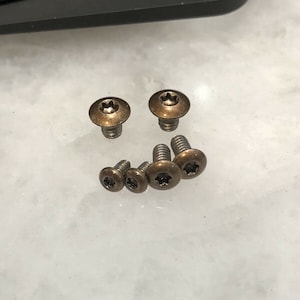 Peut inclure: Un ensemble de huit vis de couleur bronze avec une tête en forme d'étoile. Les vis sont disposées en demi-cercle sur une surface blanche.