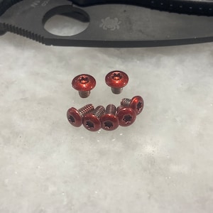May include: Close-up of eight small, red, metal screws with a star-shaped head.