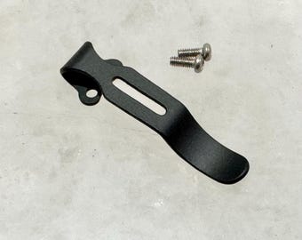 May include: Black metal pocket clip with a curved design. The clip is attached to a surface with three silver screws.