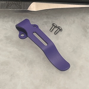 Puede incluir: Clip de bolsillo de metal morado con dos tornillos plateados. El clip está diseñado para sujetarse a una navaja de bolsillo u otra herramienta.